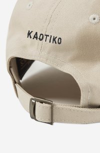 Beige-lakki, jossa on musta brodeerattu "KAOTIKO"-logo, teksturoitu kangas, säädettävä hihna ja metalliosat takana.