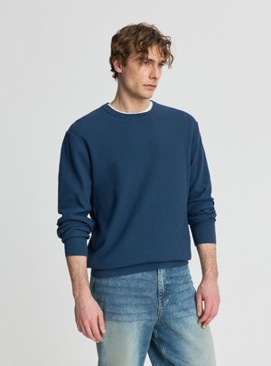 Uomo con capelli ricci che indossa un maglione blu a maniche lunghe e jeans chiari, in piedi davanti a uno sfondo grigio chiaro uniforme.