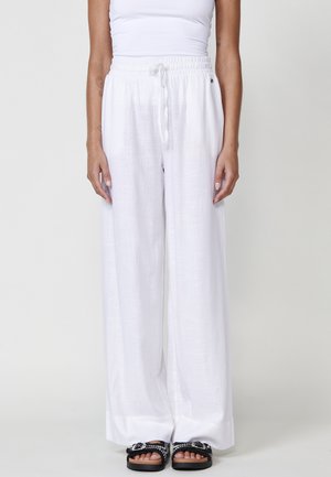 Pantaloni larghi in lino bianco con vita elasticizzata e dettaglio con cordoncino. Tessuto morbido, taglio a lunghezza completa, abbinati a sandali neri.