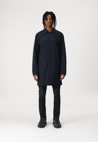Dunkelblauer, knielanger Regenmantel mit Kragen, Druckknopfverschluss, langen Ärmeln und geradem Schnitt. Getragen über schwarzen Jeans und Sneakers.