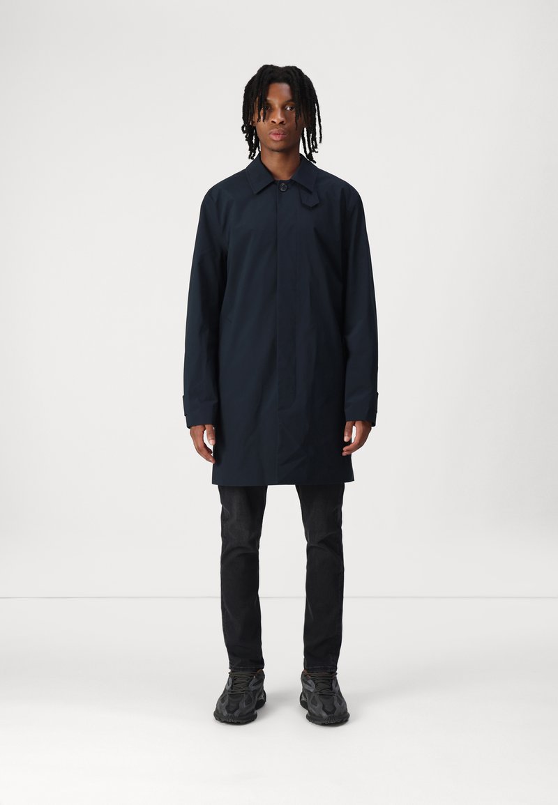 Dunkelblauer, knielanger Regenmantel mit Kragen, Druckknopfverschluss, langen Ärmeln und geradem Schnitt. Getragen über schwarzen Jeans und Sneakers.