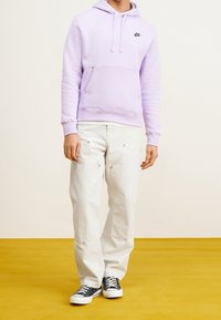 Person som bär en ljuslila Nike-huvtröja, vita arbetsklädesbyxor och svarta låga sneakers står på en guldfärg och framför en enfärgad ljus bakgrund.