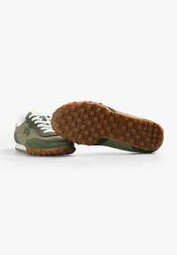 Chaussures de sport vertes avec une semelle en caoutchouc, un design à lacets et des accents blancs. Présente un fond texturé avec des motifs de grip en relief.