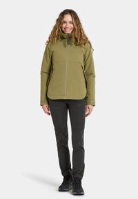 Veste softshell vert olive avec fermeture éclair complète et col montant, associée à un pantalon technique noir et des chaussures de randonnée noires.