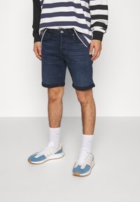 Jeansshorts i mörkblått med uppvikta ärmar, tillsammans med vita strumpor och blå sportskor med vita och orange detaljer.