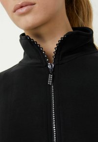 Giacca nera con zip, caratterizzata da un alto colletto, una zip decorata con dettagli trasparenti, una texture liscia e un design aderente.