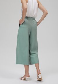 Culottes vert clair à jambes larges avec une taille élastique, comprenant deux poches arrière et une texture lisse. Portées avec des talons ouverts blancs.