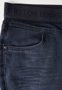 Mörkblå denimjeans med en svart elastisk midjeband som har ett präglat logotyp. Inkluderar en standard framficka och kontrasterande sömmar.