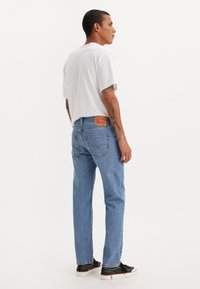 Herrecasual antrekk som inkluderer en løs hvit t-skjorte og lyseblå jeans med et lærmerke bak, kombinert med svarte sneakers.