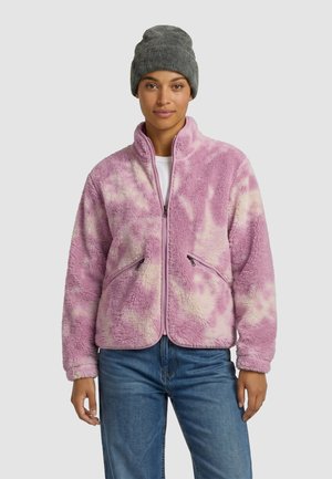 Femme portant un bonnet en laine gris, une veste polaire zippée rose et crème à motifs, une chemise blanche et un jean bleu, debout devant un fond uni.