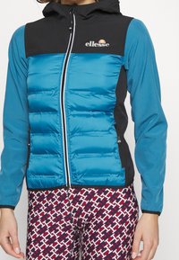 Teal puffer jacket with black accents, zip closure, hood, and logo. Features padded texture, fitted design, and side zipper pockets.