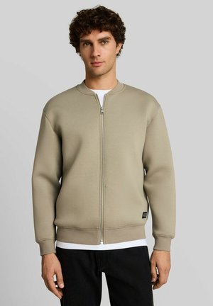 Giovane uomo con una giacca beige con cerniera sopra una camicia bianca e pantaloni neri, in piedi davanti a uno sfondo grigio chiaro uniforme.