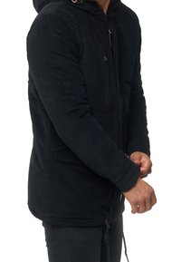 Person, der eine schwarze Cordjacke mit Kapuze trägt und den Ärmelsaum mit sichtbaren Händen anpasst, vor einem weißen Hintergrund.