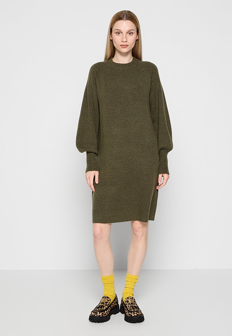 Grünes gestricktes Kleid mit lockerer Passform, langen Puffärmeln, Rundhalsausschnitt und seitlichem Schlitz, kombiniert mit gelben Socken und gemusterten Schuhen.