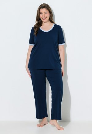 Femme souriante, debout pieds nus contre un mur blanc, portant un haut bleu marine à manches courtes et un pantalon ample assorti avec un liseré bleu clair.