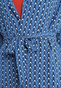 Robe bleue à motifs avec des pois blancs et des dessins de feuilles, nouée à la taille avec une ceinture en tissu assorti, portée sur un vêtement rouge.