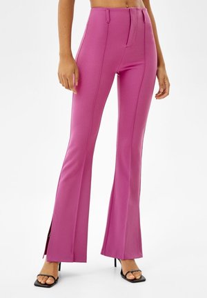 Woman wearing high-waist, pink flared pants with front seams and side slits, paired with black open-toe heeled sandals.