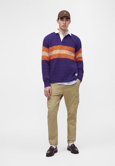 Homme portant une casquette marron, un pull violet avec des rayures orange et beige, un pantalon cargo beige, des chaussettes blanches et des mocassins marron, debout devant un fond uni.