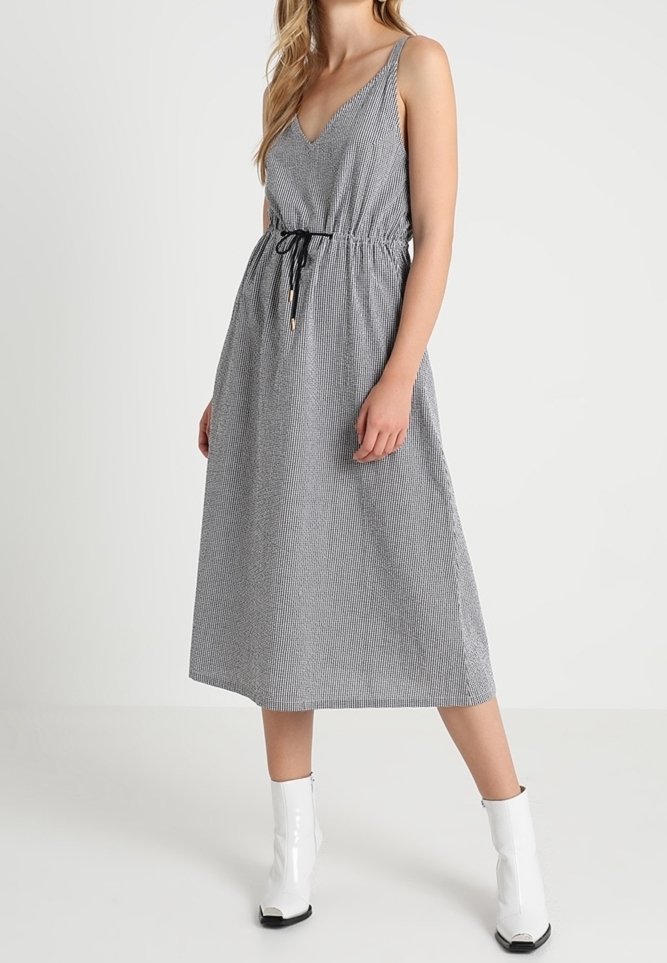 Femme portant une robe midi grise sans manches avec une taille à cordon noir et des bottines blanches, debout sur un fond uni.