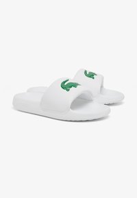Witte slide sandalen van synthetisch materiaal met een brede band met een groen krokodillenlogo en een textuur op de voetbed.