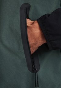 Detalle de cremallera negra en una chaqueta verde texturizada, con un bolsillo lateral diseñado para un fácil acceso a la mano. Tejido suave con un acabado mate.
