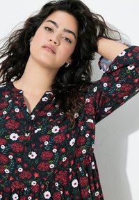 Schwarzes Blumenkleid mit roten und weißen Blumen, Knopfleiste und langen Ärmeln. Der Stoff wirkt leicht und hat eine lockere Passform.