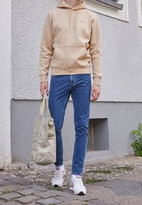 Beige mikina s kapucí, modré hubené džíny, bílé tenisky s fialovými akcenty, držící krémovou tašku, stojící na dlažební cestě.