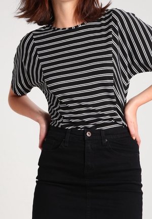 Vrouw draagt een zwarte en witte horizontaal gestreepte korte mouwen blouse, ingestopt in een zwarte rok met hoge taille, met handen in de zij.