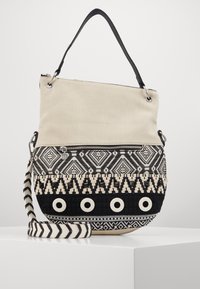 Torba na ramię z płótna i tkanej tkaniny w kolorze beżowym i czarnym. Posiada geometryczne wzory i okrągłe akcenty; zawiera regulowany paski w paski.