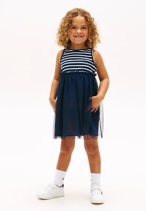 Młoda dziewczyna z kręconymi włosami, ubrana w granatową, paskowaną sukienkę bez rękawów, białe skarpetki i białe tenisówki, stojąca z rękami w kieszeniach.