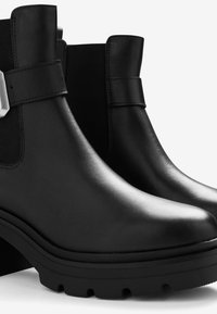 Bottines noires en cuir lisse avec un talon épais, des panneaux élastiques sur les côtés et un large détail de sangle au niveau de la cheville. Semelle texturée.