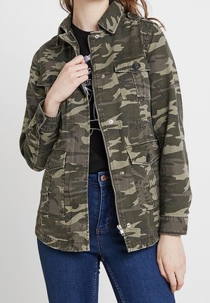Personne portant une veste camouflage verte par-dessus un top noir et un jean bleu, avec la main ajustant la veste près du col.