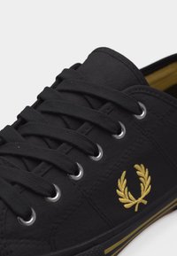 Zapatilla deportiva de lona negra con cordones negros, ojales plateados, logo de corona de laurel dorada y suela negra con rayas doradas.