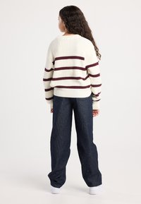 Nainen seisoo selkä kameraan, päällä vaalean beige neule, johon on kudottu burgundinpunaisia raitoja, tummat raidalliset housut ja valkoiset lenkkarit tasaisella taustalla.