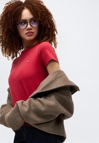 Γυναίκα με κατσαρά μαλλιά που φοράει γυαλιά με μπλε απόχρωση, κόκκινη κοντομάνικη μπλούζα και καφέ παλτό που είναι περασμένο, κοιτάζοντας πλάγια σε απλό φόντο.