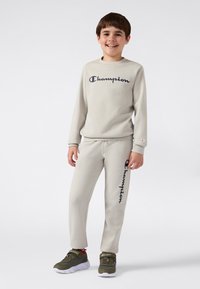 Smilende gutt som har på seg en beige Champion hettegenser og joggebukser med svart logo, står foran en hvit bakgrunn, olivengrønne sneakers synlige.