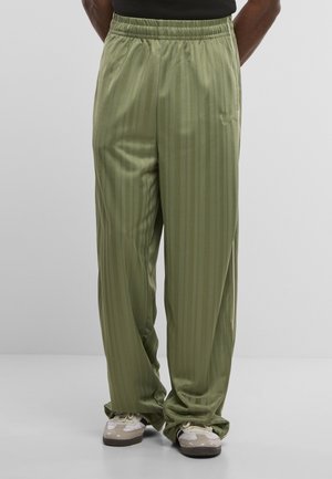 Osoba v nosí voľné olivovo zelené pásikavé nohavice a biele tenisky stojí pred jednoduchým svetlým pozadím.