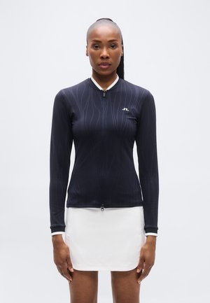 Sötétkék, hosszú ujjú, cipzáros felső fehér részletekkel, testhezálló sziluettel és finom hullámmintával. Fehér szoknyával párosítva.