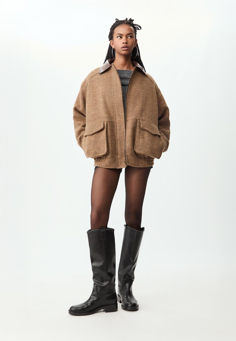 Veste en mélange de laine marron avec un col foncé, coupe oversize, deux poches avant, associée à des bottes en cuir noires jusqu'aux genoux.