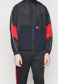 Nike Air-jacka med svart och röd design, dragkedjestängning, ståkrage, elastiska muddarna och logotyp på bröstet i vitt.
