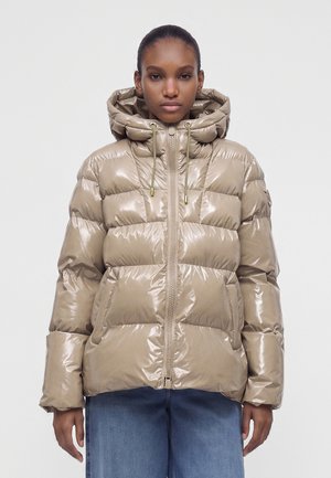 Ung kvinde iført en skinnende beige dunjakke med hætte og lynlås, kombineret med blå jeans, stående foran en ensfarvet baggrund.