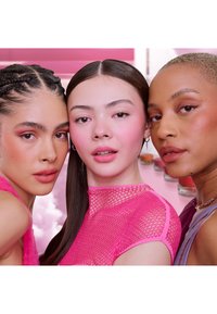 Drie diverse vrouwen met roze make-up en stijlvolle outfits die dicht bij elkaar poseren tegen een roze achtergrond met cosmetische potjes wazig op de achtergrond.