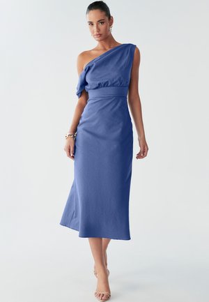 Ženska v enorazkolnem modrem obleku samozavestno pozira; njeni lasje so gladko zauzeti nazaj, nosi minimalno nakit in sandale na vezalke.