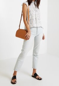 Hnědá crossbody taška zaobleného tvaru, nošená s lehkým květinovým topem a světlými modrými džíny. Vyšší černé sandály bez podpatku dotvářejí celý outfit.