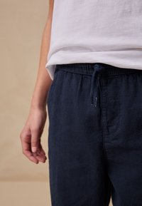 Tengerészkék színű vászonnadrág elasztikus derékpánttal és húzózsinórral, laza szabással és finom textúrával. Fehér pólóval párosítva.