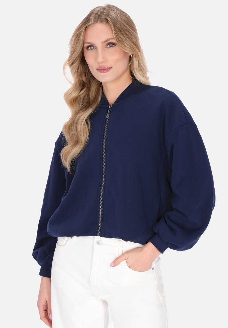 Marineblauwe bomberjack met ritssluiting en hoge kraag, met lange ballonmouwen en een zachte textuur, gecombineerd met witte broek.