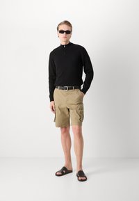 Tricou negru cu fermoar și mâneci lungi, asortat cu pantaloni scurți cargo bej. Cureaua neagră și sandalele tip slide completează ținuta. Design simplu și funcțional.