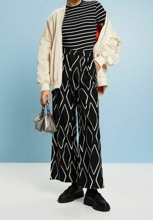 Persona con camiseta de rayas blancas y negras, chaqueta beige de gran tamaño, pantalones negros de pierna ancha con patrón geométrico blanco, zapatos negros, sosteniendo una pequeña bolsa plateada.