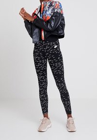 Černá zkrácená bunda s barevným abstraktním vzorem; černé legíny s bílým potiskem loga Nike; b éžové tenisky s texturovanými detaily.