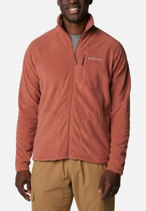 Uomo che indossa una giacca in pile Columbia color ruggine con cerniera, abbinata a pantaloni cargo beige, sorridente davanti a uno sfondo semplice.